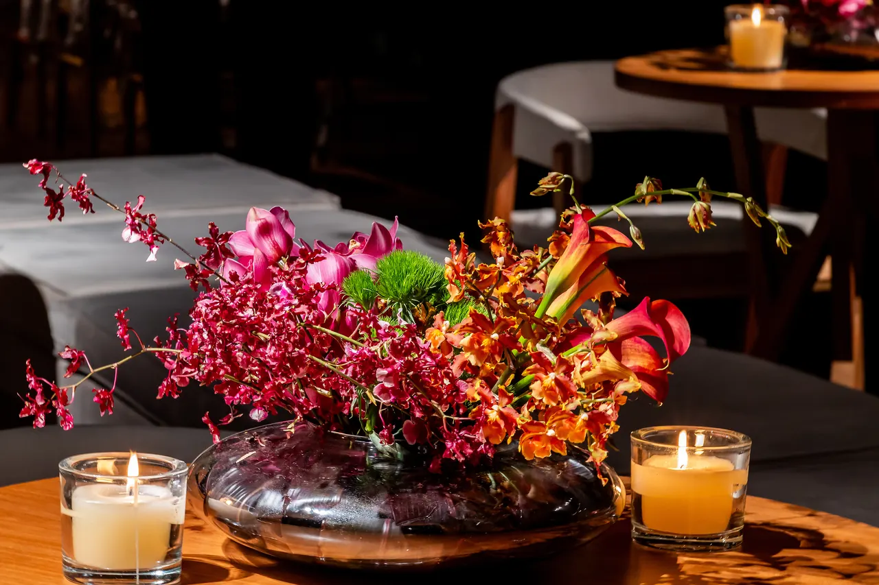 Arranjo de flores magenta e laranja entre velas acesas, sobre uma mesa
